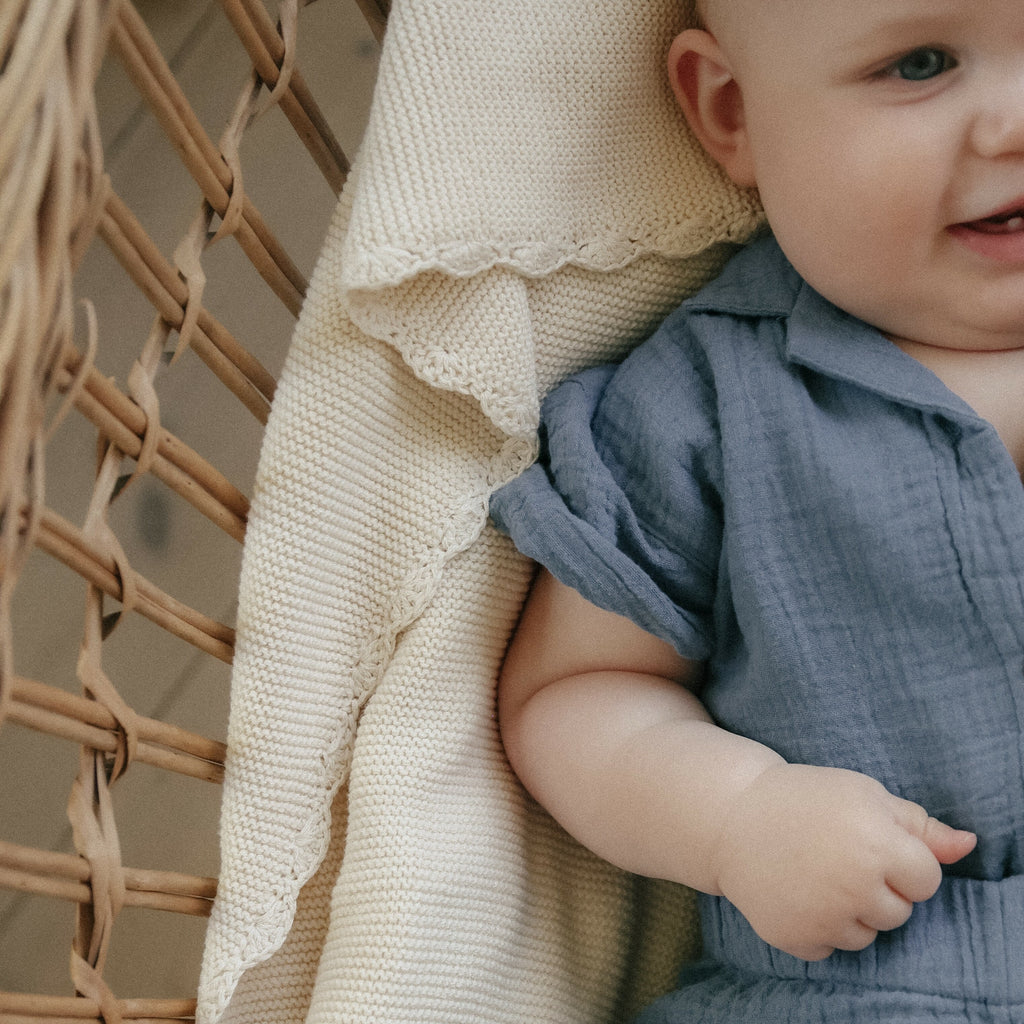 Cotton Knit Baby Blanket in Ivory is displayed under a baby boy in a woven bassinet.